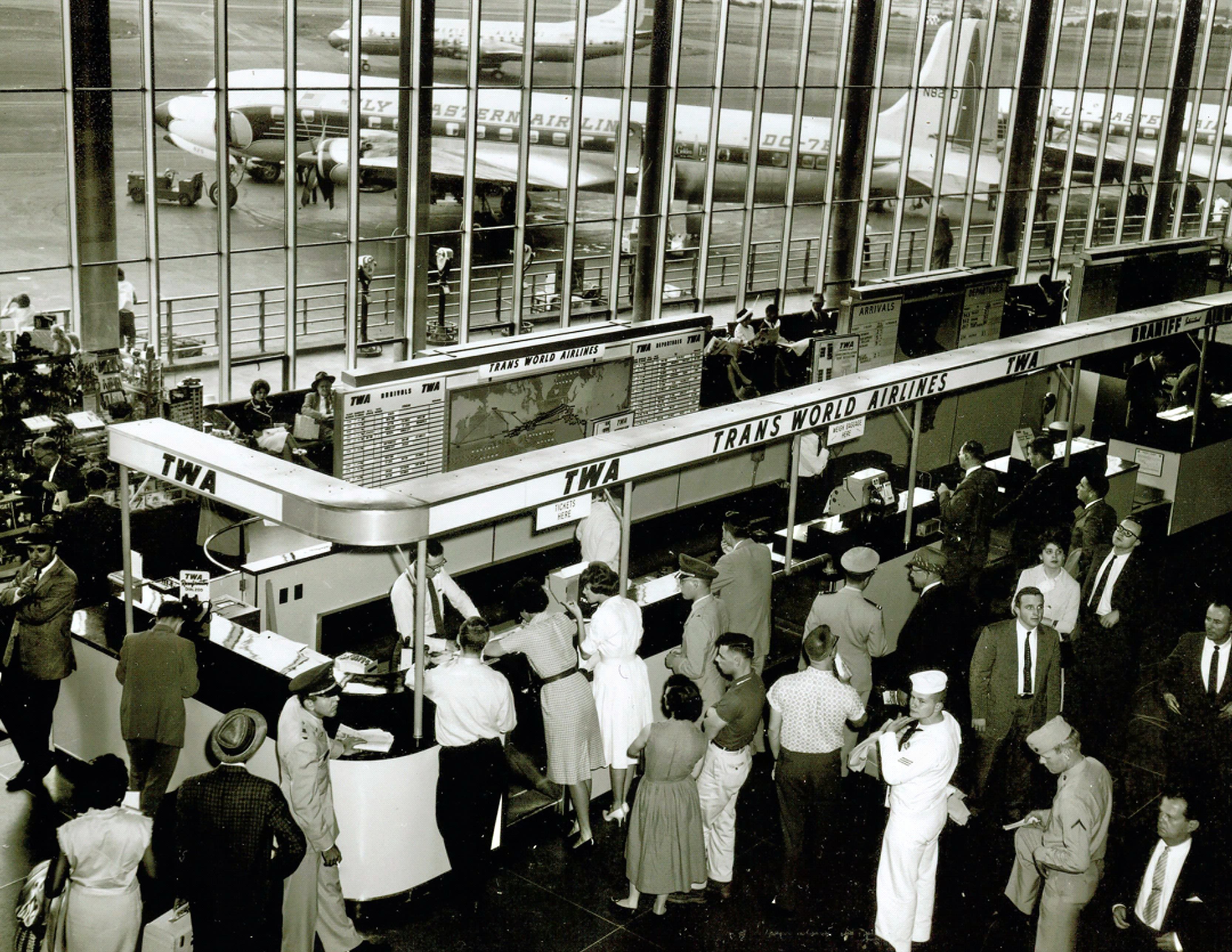 1960s passengers at the airport terminal showing crowded TWA ticket counter and Eastern Air Lines DC-7 (Wikimedia Commons: FAA photo).